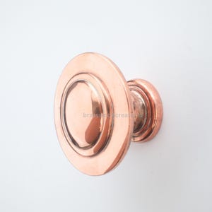 May include: A copper-coloured cabinet knob with a circular design. The knob has a flat, round face with a raised, concentric ring around a central, smaller circle. The stem is also copper-coloured.