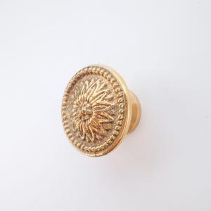 Op de afbeelding: Een goudkleurige kastknop met een zonnebloemontwerp. De knop heeft een ronde voorkant met een gedetailleerde zonnebloem in het midden, omgeven door een kralenrand. De hardware is van metaal.