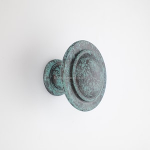 May include: A weathered, round door knob with a distressed teal and bronze finish. The knob has a circular design with concentric rings, suggesting a vintage or antique style. The hardware is mounted on a white surface.