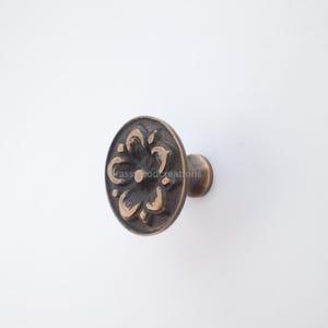 May include: Bronze cabinet knob with a floral design. The knob has a circular face with an embossed flower pattern and a short, cylindrical stem. The hardware is set against a white background.