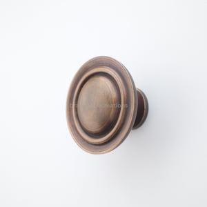 Puede incluir: Un pomo de armario de color bronce con un diseño circular. El pomo presenta un círculo central ligeramente elevado rodeado de anillos concéntricos. Este herraje decorativo es probablemente para muebles o armarios.