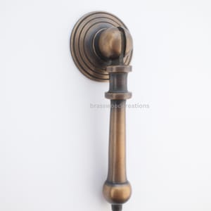 May include: Antique bronze cabinet pull with a round backplate and a decorative handle. The handle has a unique, elongated shape with a rounded end. The pull is mounted on a white surface. The text "brasswood creations" is visible.