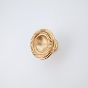 May include: A gold-colored cabinet knob with a circular design. The knob features a detailed, ridged outer ring and a rounded center. The hardware is made of metal and is designed for furniture. The knob is set against a white background.