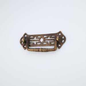 Puede incluir: Un tirador de cajón de latón con un diseño geométrico decorativo. El tirador tiene una forma rectangular con un patrón recortado y una pequeña manija curva.