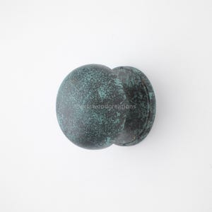May include: A weathered, round door knob with a mottled teal and dark green finish. The knob has a circular shape with a smaller, attached circular base. The surface has a textured appearance, suggesting an aged or antique aesthetic.