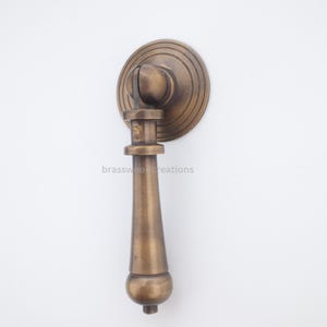 May include: A bronze-colored cabinet pull with a circular backplate and a teardrop-shaped handle. The handle is suspended from a cylindrical post. The text "brasswood creations" is printed on the handle.
