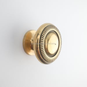 May include: A brass door knob with a circular design. The knob has a round, ridged outer ring and a smooth, central disc. The hardware is a warm, golden color, and is set against a white background. The knob is likely for a door or cabinet.