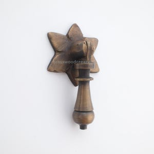 Op de afbeelding: Een bronzen trekker met een stervormige achterplaat. De trekker heeft een decoratieve, langwerpige vorm met een afgeronde basis. De tekst "brasswoodcreation" is zichtbaar op de achterplaat. Deze hardware is geschikt voor kasten en laden.