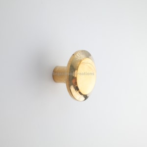May include: A gold cabinet knob with an oval face and a cylindrical base. The knob has a polished finish and is set against a white background. The text "brasswood creations" is printed on the knob.