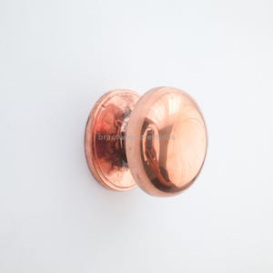 May include: A copper-colored door knob with a round, polished handle and a circular backplate. The hardware is mounted on a white surface, reflecting light. The knob has a shiny, reflective surface.