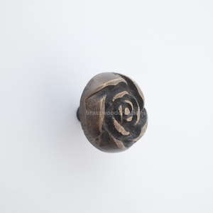 Könnte beinhalten: Schrankknopf in Bronzefarbe mit Rosenmuster. Die Rose hat detaillierte Blütenblätter und ist mittig auf dem Knopf angeordnet. Der Knopf ist mit einer schwarzen Schraube befestigt.