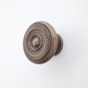 Könnte beinhalten: Ein bronzefarbener Schrankknauf mit kreisförmigem Design. Der Knauf hat konzentrische Kreise und ein detailliertes Muster am Rand. Der Text "brasswood creations" ist sichtbar.