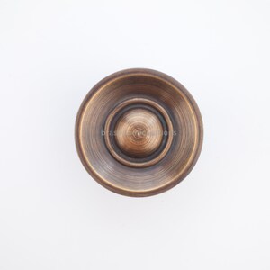 Puede incluir: Un pomo de armario de color bronce con un dise&ntilde;o circular. El pomo tiene una espiral central elevada rodeada de anillos conc&eacute;ntricos. La palabra "brass" es visible en el pomo. El pomo est&aacute; sobre un fondo blanco.