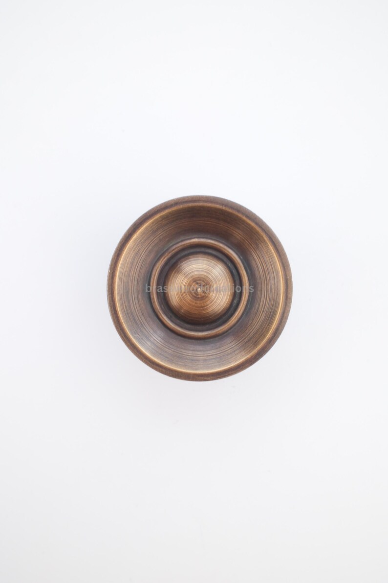 Puede incluir: Un pomo de armario de color bronce con un dise&ntilde;o circular. El pomo tiene una espiral central elevada rodeada de anillos conc&eacute;ntricos. La palabra "brass" es visible en el pomo. El pomo est&aacute; sobre un fondo blanco.