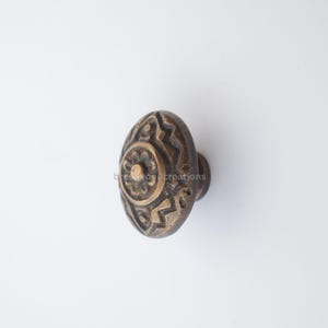May include: An antique-style brass cabinet knob with an oval shape. The knob features an intricate, embossed design with a central floral motif and geometric patterns. The knob is a warm, aged bronze color, suggesting a vintage aesthetic.