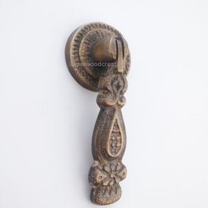 May include: Ornate bronze-colored drawer pull with a circular backplate and a decorative handle. The handle features floral and teardrop-shaped details. The hardware is designed for furniture and cabinetry.