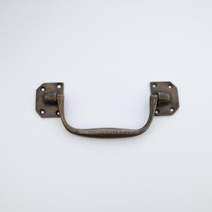 Op de afbeelding: Een messing kastgreep met een afgeronde rechthoekige vorm. De greep heeft een gebogen, gladde stang en twee vierkante platen met elk vier gaten.