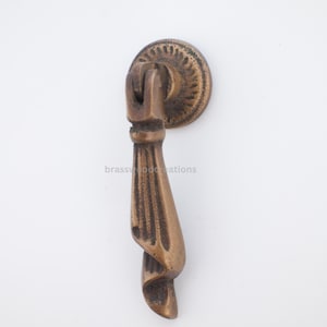 Puede incluir: Un tirador de armario de color bronce con un diseño decorativo. El tirador presenta una placa trasera redonda con un patrón detallado y un asa en forma de lágrima. El texto "brasswoodcreations" es visible.