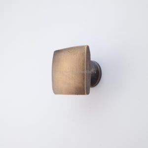 Op de afbeelding: Een vierkante, antieke messing kastknop met afgeronde randen. De knop heeft een donkere, cirkelvormige basis en een getextureerd oppervlak. De hardware is ontworpen voor meubels en woondecoratie.