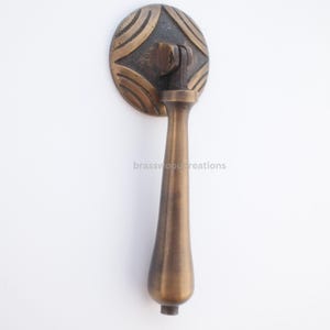 Peut inclure: Poignée de tiroir en laiton antique avec une plaque arrière circulaire décorative ornée d'un motif géométrique en relief. La poignée a une forme de goutte d'eau et est fixée à la plaque arrière par un petit support. Le texte "brasswood creations" est imprimé.