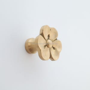 Puede incluir: Un tirador de armario con forma de flor de latón. El tirador presenta un diseño floral detallado con cinco pétalos y un capullo central. El herraje está montado sobre una superficie blanca, mostrando el acabado metálico cálido.