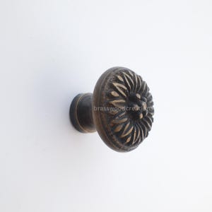 May include: Bronze-toned cabinet knob with a floral design. The knob has a round, textured face with a detailed pattern and a cylindrical base. The hardware is set against a white background.