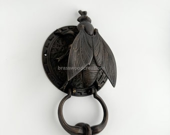 May include: A black metal door knocker shaped like a fly with intricate details. The knocker is attached to a round base with a decorative border. The fly has a raised design and is positioned with its wings spread out.