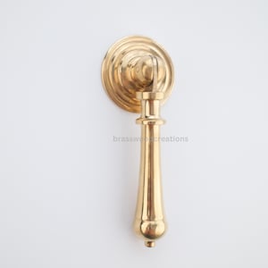 Puede incluir: Un tirador de armario dorado con una placa trasera redonda y un mango largo y elegante. El mango tiene un extremo bulboso y un cuerpo cilíndrico. El texto "brasswoodcreations" es visible en el mango. Adecuado para la restauración de muebles.