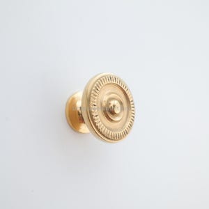 May include: A gold-colored cabinet knob with a circular design. The knob features concentric circles and a central raised button. The outer ring has a detailed, textured pattern. The hardware is mounted on a white background.