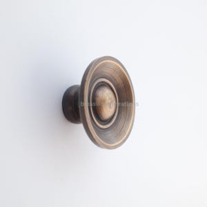 May include: A bronze-toned cabinet knob with a circular design. The knob features concentric rings and a central, smaller circle. The hardware is mounted on a dark, cylindrical base, suitable for furniture or cabinetry.