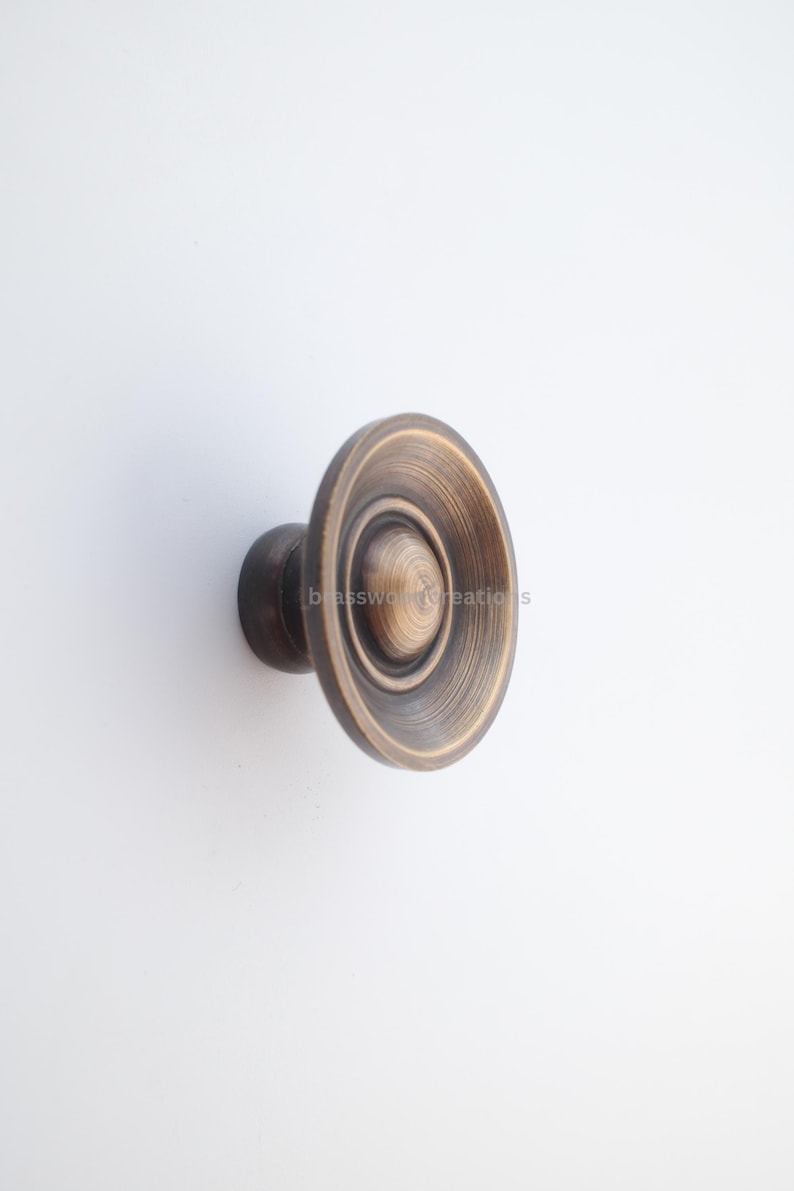 Puede incluir: Un pomo de armario de color bronce con un dise&ntilde;o circular. El pomo presenta anillos conc&eacute;ntricos y un c&iacute;rculo central m&aacute;s peque&ntilde;o. El herraje est&aacute; montado sobre una base cil&iacute;ndrica oscura, adecuado para muebles o armarios.