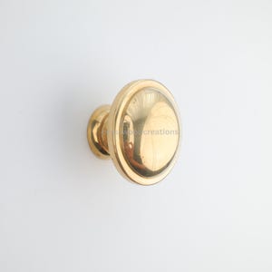 May include: A gold-colored cabinet knob with a rounded, disc-shaped handle. The knob has a polished finish and a small, circular base. The hardware is set against a white background.