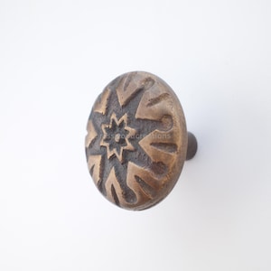 Może przedstawiać: Mosiężny uchwyt do szafki z motywem gwiazdy. Gałka ma okrągły kształt ze szczegółowym, teksturowanym wzorem. Gwiazda jest wyśrodkowana, otoczona stylizowanymi kształtami. Gałka jest zamontowana na białym tle.