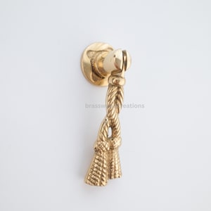 May include: A gold-colored cabinet pull with a rope design. The pull features a circular base, a decorative rope-like handle, and a tassel-like end. The hardware is made of brass and is suitable for furniture restoration or new builds.