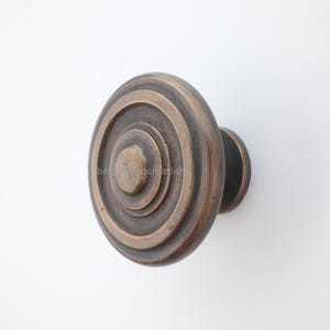 Könnte beinhalten: Ein bronzefarbener Schrankknauf mit kreisförmigem Design. Der Knauf weist konzentrische Ringe auf, die einen geschichteten Effekt erzeugen. Die Hardware ist auf einer kleinen, runden Basis montiert. Die Gesamtästhetik ist vintage und dekorativ.