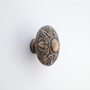 Op de afbeelding: Bronzen kastknop met een ovaal, sierlijk ontwerp. De knop heeft een centraal cirkelvormig element omgeven door wervelende, gestileerde bloempatronen. De hardware is waarschijnlijk voor meubels of kasten.