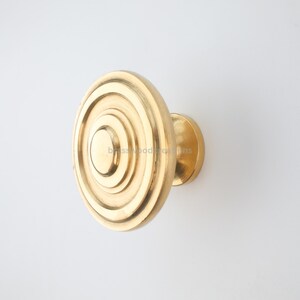 Puede incluir: Un pomo de armario dorado con un diseño circular. El pomo presenta anillos concéntricos, creando un efecto de capas. El herraje es probablemente de latón y está diseñado para muebles o armarios.