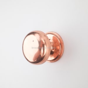 May include: A pair of copper-colored door knobs. The knobs are round with a polished finish, reflecting light. The base of each knob is a flat, circular plate. The text "brasswood creations" is visible on the surface.