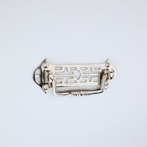 Könnte beinhalten: Ein silberner Schubladenzug mit einem dekorativen, geometrischen Design und einem gebogenen Griff.