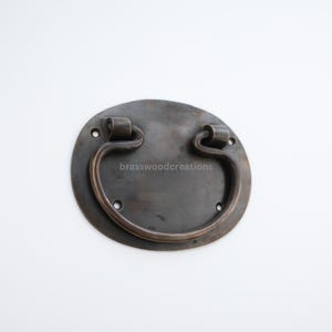 May include: A dark brown metal door pull with a rounded oval shape and a curved handle. The handle is attached to the oval plate with two small screws.