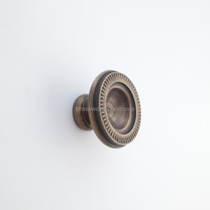 May include: A bronze-colored cabinet knob with a circular design. The knob features a detailed, textured pattern around the outer edge and a recessed center. The hardware is likely for furniture or cabinetry.