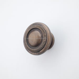 Könnte beinhalten: Bronzefarbener Schrankknauf mit kreisförmigem Design. Der Knauf hat eine zentrale, leicht erhabene Scheibe, die von einem detaillierten, strukturierten Ring umgeben ist. Der äußere Rand hat kleine, dreieckige Muster.