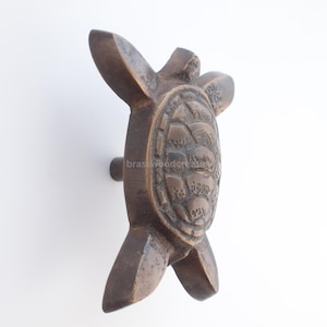 May include: Bronze-colored turtle-shaped cabinet pull. The pull features a detailed turtle design with a textured shell and flipper-like arms and legs. The pull is mounted on a small, round base.