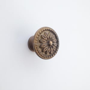 Puede incluir: Un pomo de armario de color bronce con un diseño floral. El pomo tiene una cara circular con un patrón en relieve de pétalos y un borde con cuentas. El vástago es cilíndrico. El pomo es probablemente de metal.