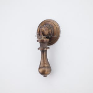May include: A bronze-colored cabinet pull with a round backplate and a decorative handle. The handle has a bulbous shape and a detailed design. The text "brasswoodcreations" is visible on the backplate.