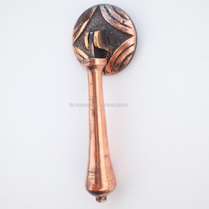 May include: Copper-colored cabinet pull with a decorative circular backplate featuring a textured pattern. The handle has a long, tapered design. The text "brasswoodcreations" is printed on the handle.
