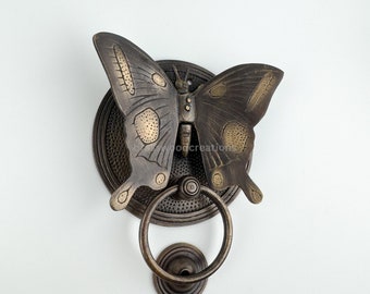 Könnte beinhalten: Ein Türklopfer aus Messing in Form eines Schmetterlings mit goldenen Akzenten. Der Schmetterling ist an einer runden Messingplatte mit einem Ring befestigt.