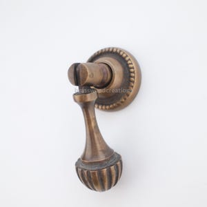 May include: Antique bronze cabinet pull with a decorative, ribbed bottom and a round backplate. The pull has a classic design and is likely made of metal. The hardware is a warm, aged bronze color.