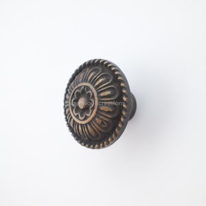 May include: Bronze-toned cabinet knob with a floral design. The knob features a central flower surrounded by ornate, curved patterns and a rope-like border. A classic hardware piece for furniture.