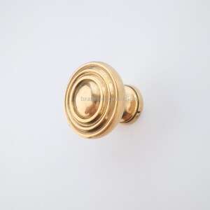 Könnte beinhalten: Ein goldfarbener, runder Türknauf mit einem dekorativen, konzentrischen Ringdesign. Der Knauf hat eine kleine, kreisförmige Basis und eine glatte, polierte Oberfläche. Das Produkt ist wahrscheinlich für die Inneneinrichtung.